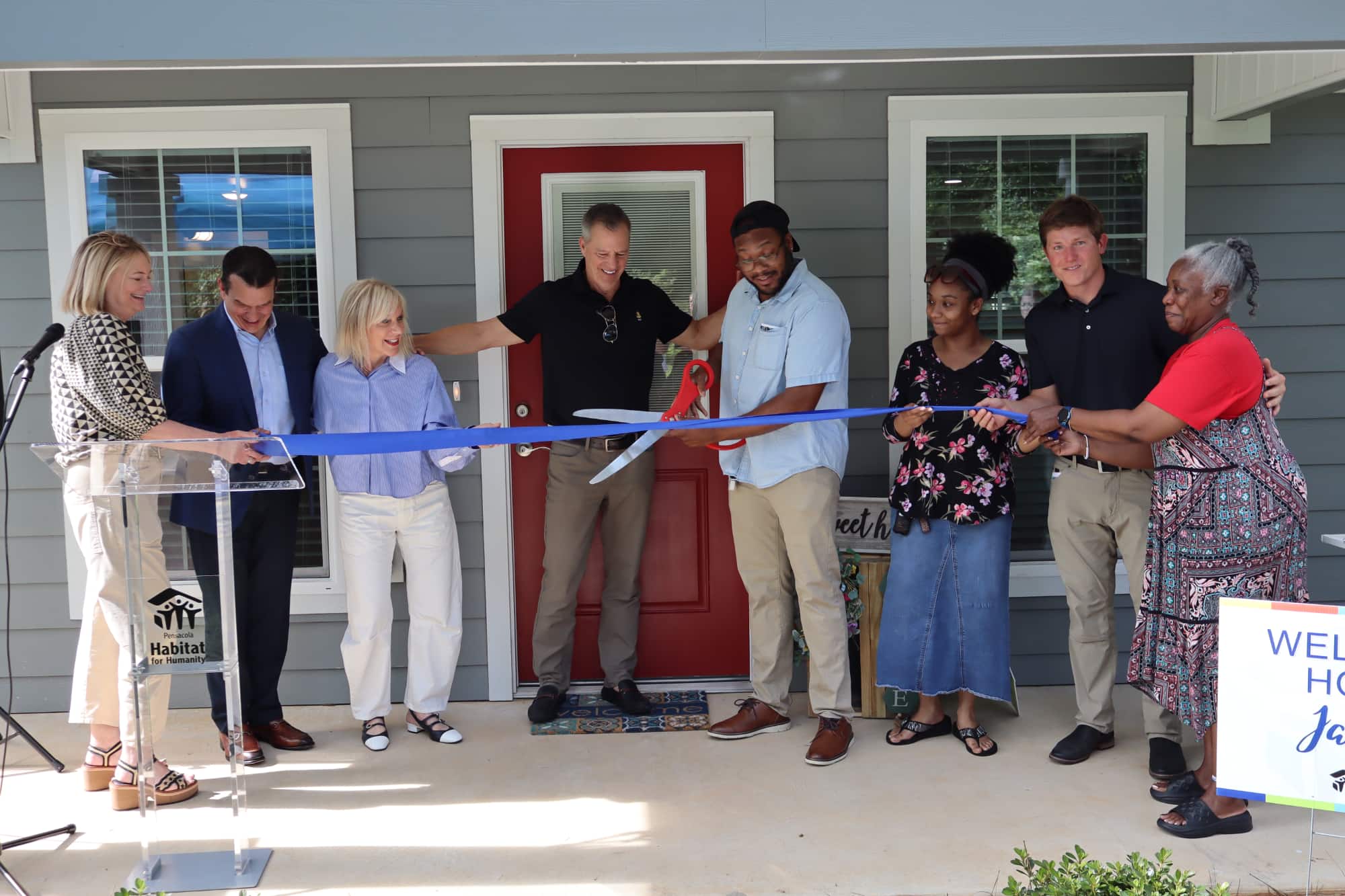 A group of people on the porch of a house using giant scissors to cut a blue ribbon.